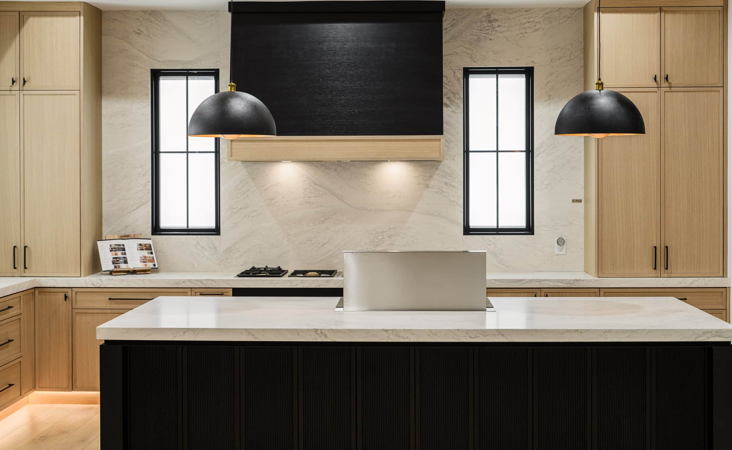 A black ventilation hood over a stone backsplash in the Roseville showroom, a gas cooktop and a downdraft vent on the island in front, light oak cabinetry either side
