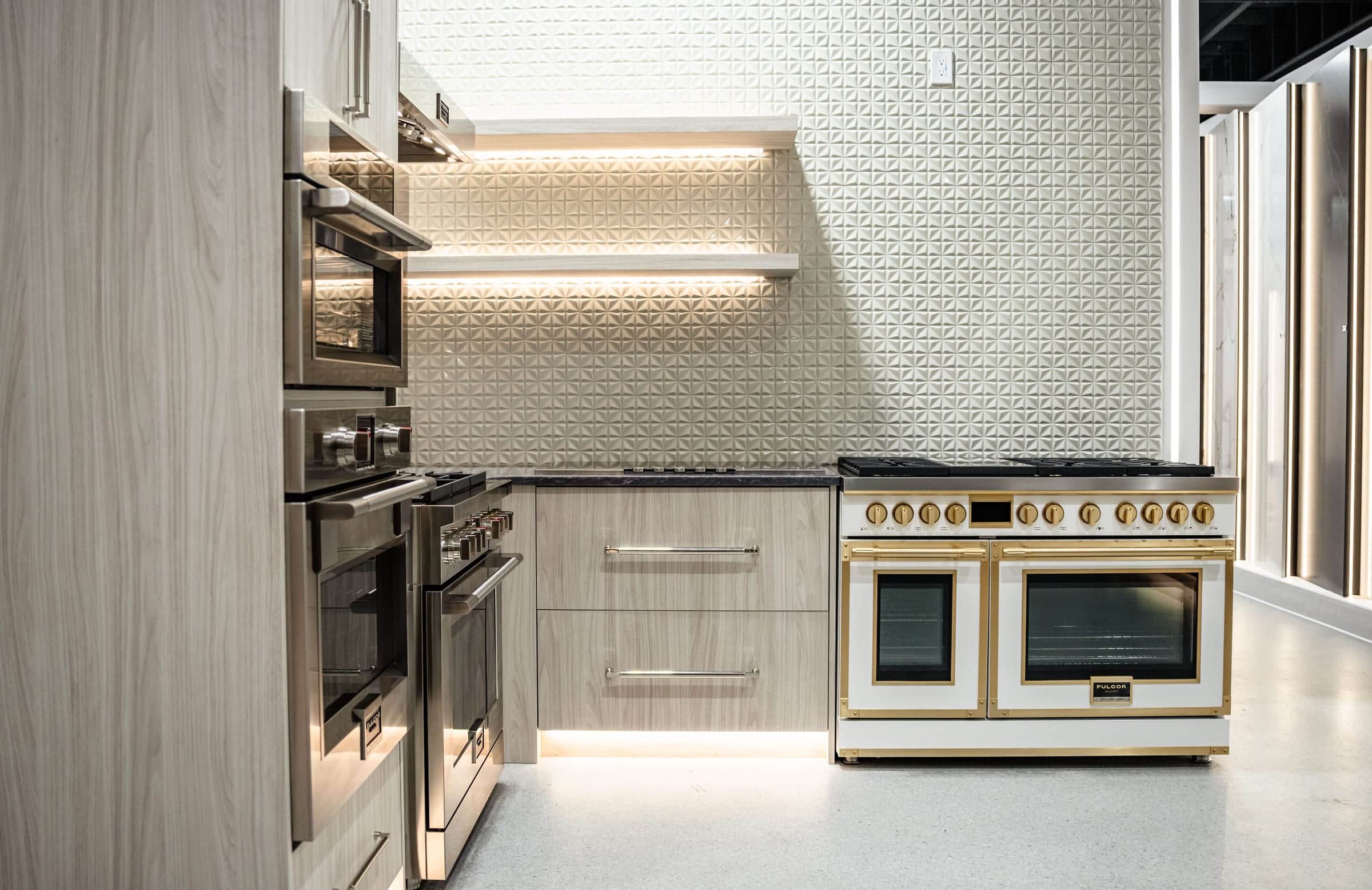 A Roseville showroom display with a pale oak tall wall of two wall ovens, a stainless range and a white range with brass trim under lit floating shelves on a pleated white tile wall