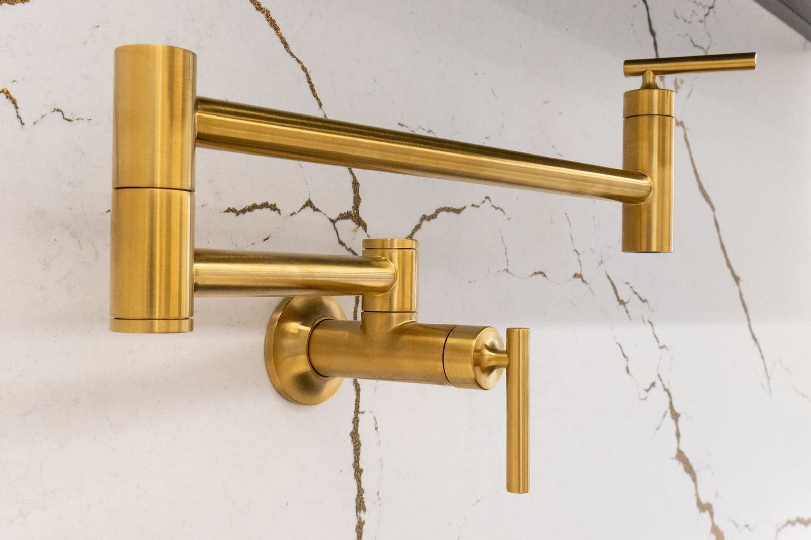 A brushed brass wall-mounted pot filler with a folding arm on a veined white stone backsplash