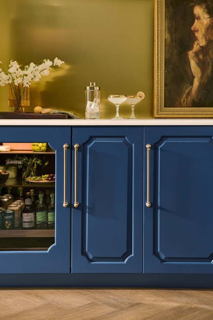 A Sub-Zero glass-door undercounter refrigerator stocked with drinks and produce beside a closed cabinet, set into deep blue bar cabinetry with a brass wall behind
