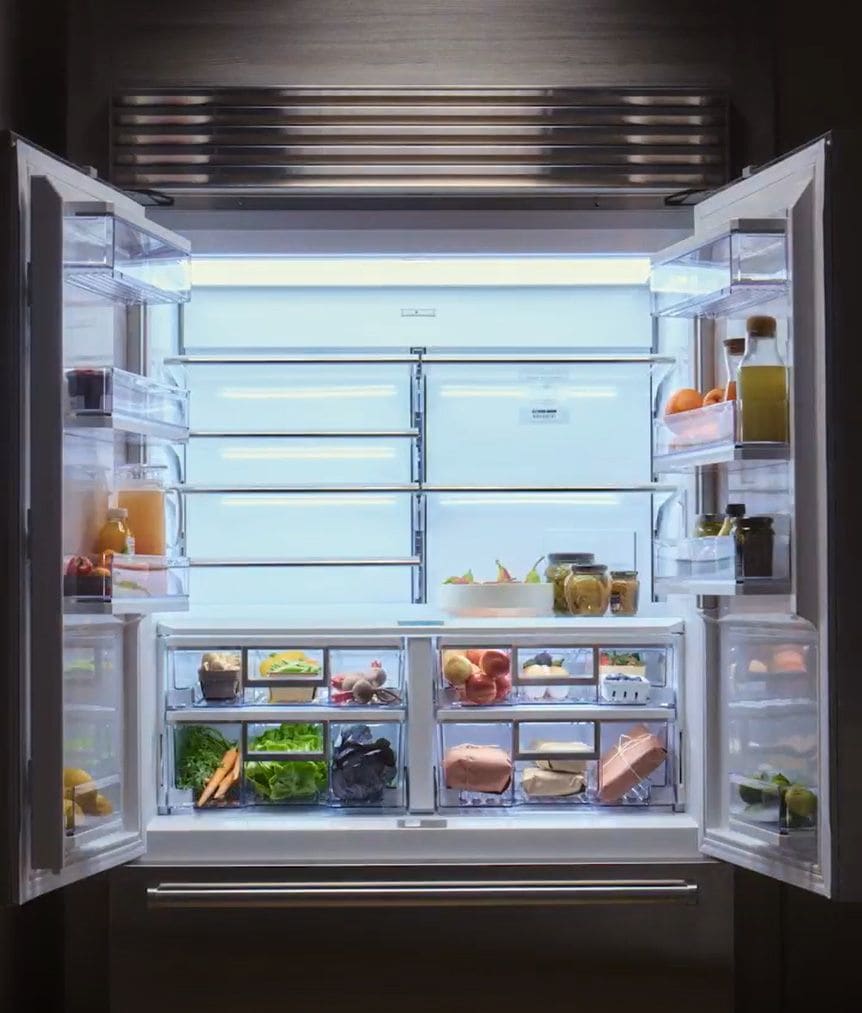 Built-in Sub-Zero refrigerator with both French doors open, showing lit glass shelves and drawers stocked with fresh produce, dairy, and preserves