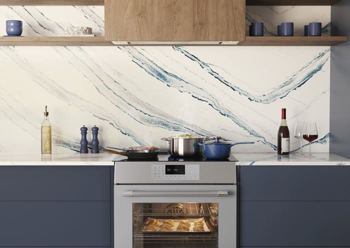 Front-on view of a Thermador range and wall hood in a navy kitchen with a large blue-veined marble slab backsplash and pastries baking in the open oven